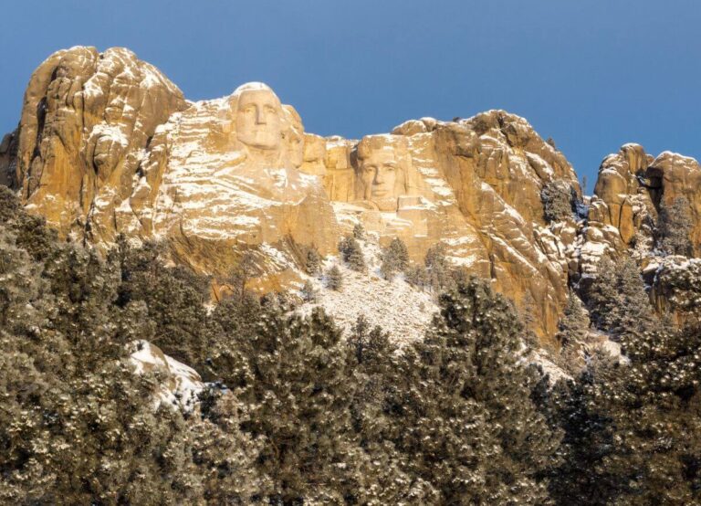 Mount Rushmore National Memorial, with the carved faces of George Washington and Thomas Jefferson partly covered in snow, surrounded by rugged granite cliffs. Evergreen trees dusted with snow fill the foreground beneath a clear blue sky.