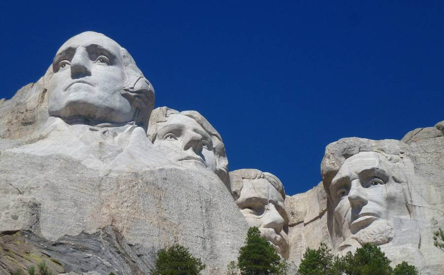 Close-up view of Mount Rushmore National Memorial under a clear blue sky, featuring the massive granite carvings of four U.S. presidents, George Washington, Thomas Jefferson, Theodore Roosevelt, and Abraham Lincoln. The white stone faces stand out against the vivid sky, with green pine trees scattered at the base of the monument.