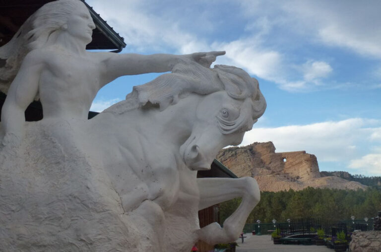 A white statue model of the Crazy Horse Memorial in the foreground, showing the Native American leader riding a horse and pointing forward. Behind the statue, the actual Crazy Horse mountain carving can be seen in the distance, partially completed with the face finished and a tunnel carved beneath the arm. The setting includes a paved viewing area with potted plants and trees below the mountain, under a partly cloudy sky.
