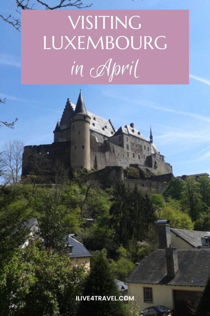a pinterest pin about visiting luxembourg in april showing a castle stood on top of a hill with grass and trees coming down the hill and houses at the bottom on a sunny day with blue skies