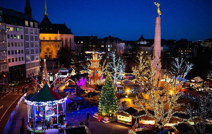 a christmas market with buildings to the left, trees with lights, a christmas tree and some stalls
