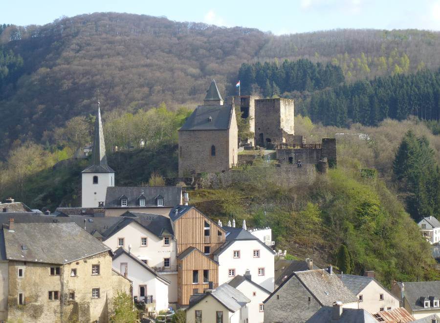 the ruins of a castle sat on a hill just above a town with the houses below, the area is surrounded by trees