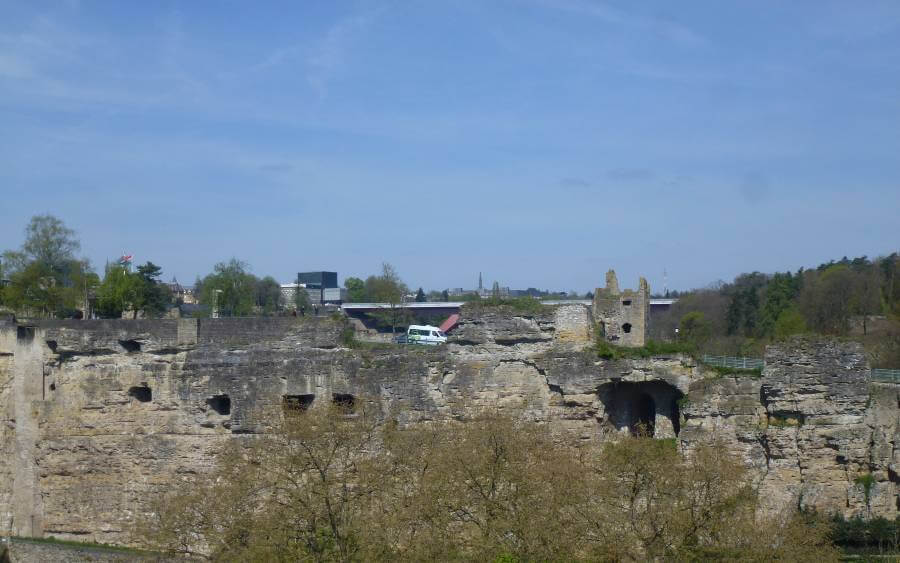 a rock carved to make a wall with square holes in it, with ruins behind it and some trees in the background