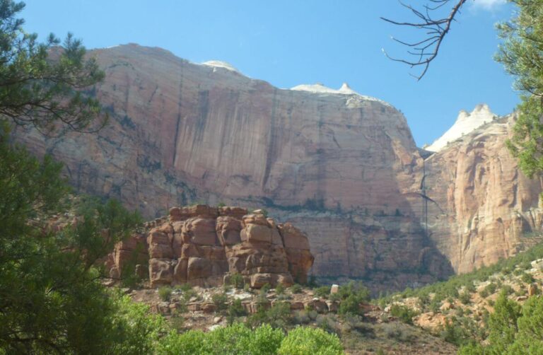 a valley with trees and steep cliffs on the side, the cliffs are a mixture of orange and grey on a clear day with blue skies