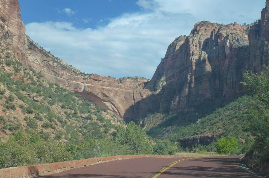 a road winding through a valley, with trees and bushes and large steep cliffs and what looks like it used to be a waterfall coming down over one of the cliffs