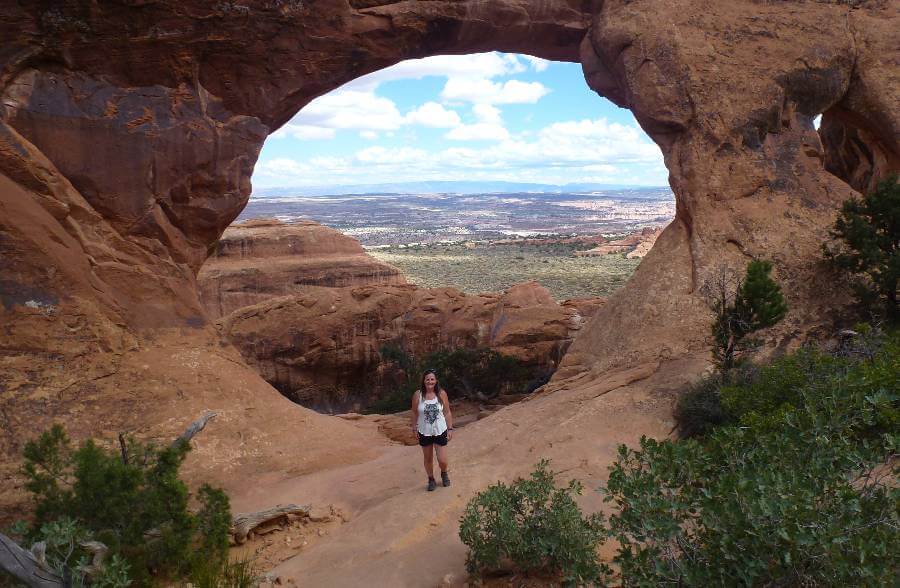 a girl in a white tshirt stood before a large arch with a valley in the background through the arch