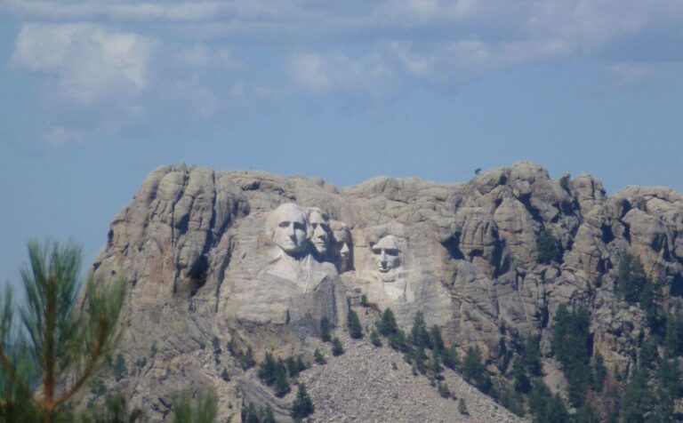 Distant view of Mount Rushmore in South Dakota, with the carved faces of four U.S. presidents, George Washington, Thomas Jefferson, Theodore Roosevelt, and Abraham Lincoln, set against rugged granite cliffs. The monument is framed by green pine trees in the foreground under a bright blue sky with scattered white clouds.