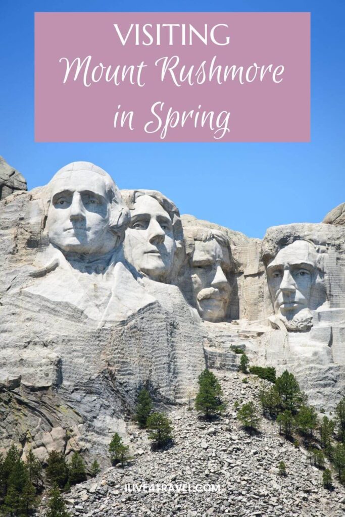 Pinterest pin featuring Mount Rushmore National Memorial in South Dakota, with the colossal stone carvings of George Washington, Thomas Jefferson, Theodore Roosevelt, and Abraham Lincoln standing out in bright spring sunlight against a clear blue sky. The granite cliff is surrounded by scattered evergreens and rocky slopes. A text box at the top reads, “Visiting Mount Rushmore in Spring”