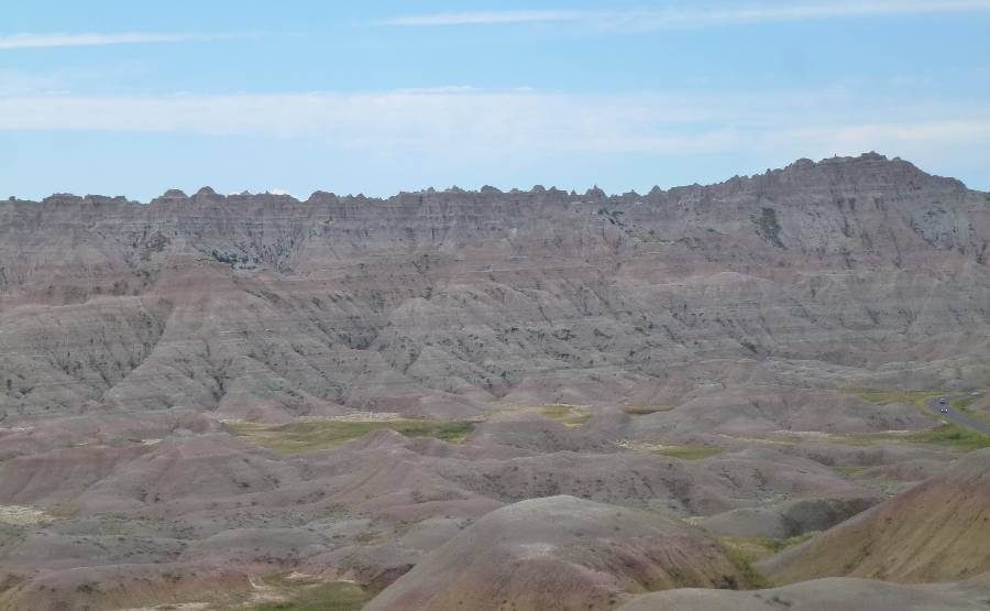 Badlands National Park in South Dakota, featuring rolling, eroded hills with layered bands of tan, pink, and gray rock, sparse vegetation in the valleys, and jagged ridgelines stretching across the horizon under a blue sky