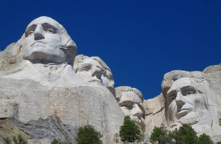Close-up view of Mount Rushmore in South Dakota, showing the massive granite faces of George Washington, Thomas Jefferson, Theodore Roosevelt, and Abraham Lincoln carved into the mountainside. The pale stone sculptures rise above evergreen trees under a deep blue summer sky.