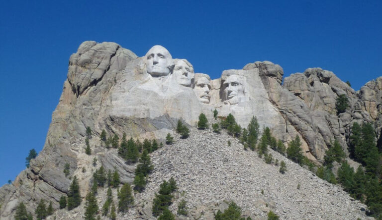 Mount Rushmore National Memorial featuring the carved granite faces of four U.S. presidents—George Washington, Thomas Jefferson, Theodore Roosevelt, and Abraham Lincoln—set against a clear blue sky, with pine trees scattered below the sculpture.