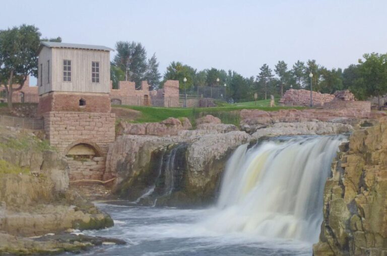 Wide view of a cascading waterfall at Falls Park in Sioux Falls, South Dakota, with white water flowing over layered rock ledges into a river below. Historic stone ruins and a small wooden mill building sit along the cliff edge, surrounded by green grass and leafy trees under a pale summer sky.