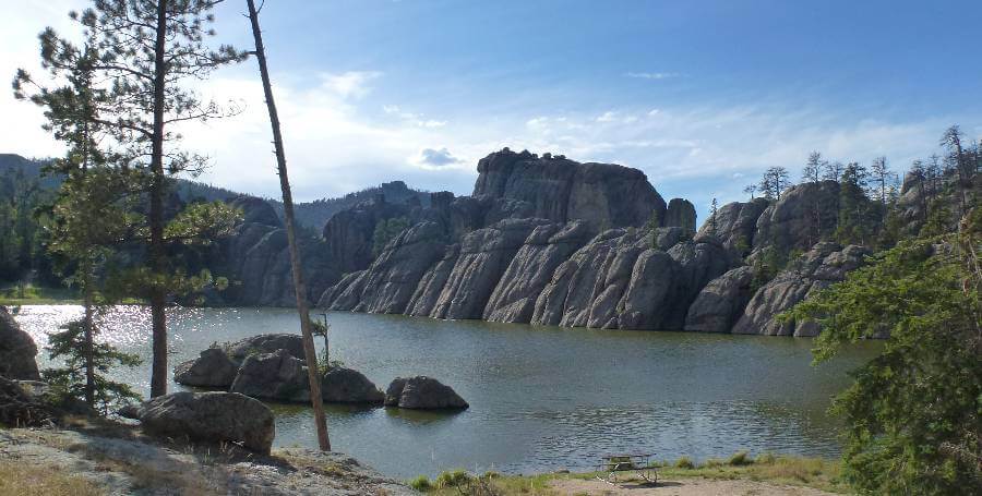 Scenic view of a calm lake in South Dakota surrounded by rugged granite rock formations and scattered pine trees. Large stone outcrops rise along the shoreline, reflected in the water, while evergreen trees frame the scene beneath a bright blue sky, capturing a peaceful spring landscape.