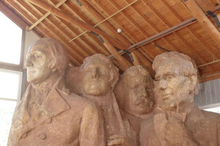 Plaster model of Mount Rushmore displayed indoors, showing detailed busts of George Washington, Thomas Jefferson, Theodore Roosevelt, and Abraham Lincoln. The figures are sculpted in brown plaster with visible textures and are housed under a wooden-beamed roof with natural light filtering through the windows.
