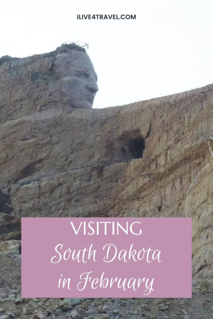 Pinterest pin featuring the Crazy Horse Memorial in South Dakota, showing the massive stone-carved face emerging from a rugged granite mountainside. A soft pink text overlay reads “Visiting South Dakota in February”