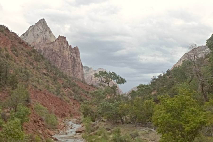 a river flowing through a canyon with orange sloping sides and some white mountains in the near distance with trees and bushes to the side of the river