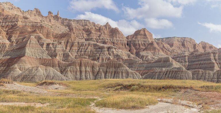View of Badlands National Park in South Dakota, featuring sharply eroded rock formations with layered bands of tan, red, and gray stone. Rolling grassy patches fill the foreground, while jagged ridges and spires rise dramatically under a partly cloudy sky, showcasing the rugged terrain of the Badlands in spring.