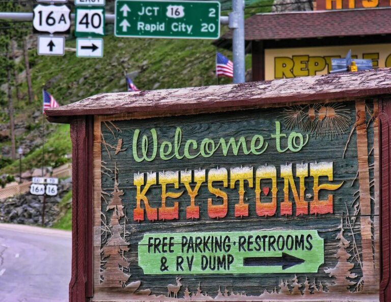 Wooden roadside sign reading “Welcome to Keystone” with colorful carved letters and illustrations of trees and fireworks. Below, a smaller sign offers “Free Parking, Restrooms & RV Dump” with an arrow. In the background are highway signs pointing toward Rapid City and U.S. Routes 16 and 40, along with American flags.