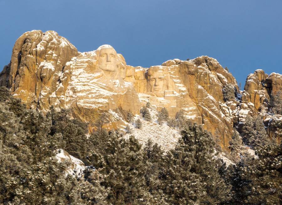 Mount Rushmore appears in its winter setting, with the towering granite sculptures carved into warm-toned stone and lightly covered in snow. Frosted evergreen trees blanket the landscape below, while the landmark rises clearly above the forest under a crisp blue sky