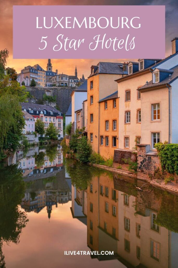 A Pinterest pin titled 'Luxembourg 5 Star Hotels' in white text on a mauve banner. The background photo captures a breathtaking golden sunset view of the Grund district in Luxembourg City, with a row of tall, centuries-old buildings in warm ochre, peach, and cream tones lining the bank of the Alzette River. Their colorful facades and the dramatic orange and purple-tinged sky are perfectly mirrored in the glassy, still water below. High above on the clifftop to the left, the historic old town fortifications and buildings, including a church spire, rise dramatically against the moody dusk sky. Lush green weeping willows and vegetation frame the left bank, adding to the romantic, painterly atmosphere of the scene.