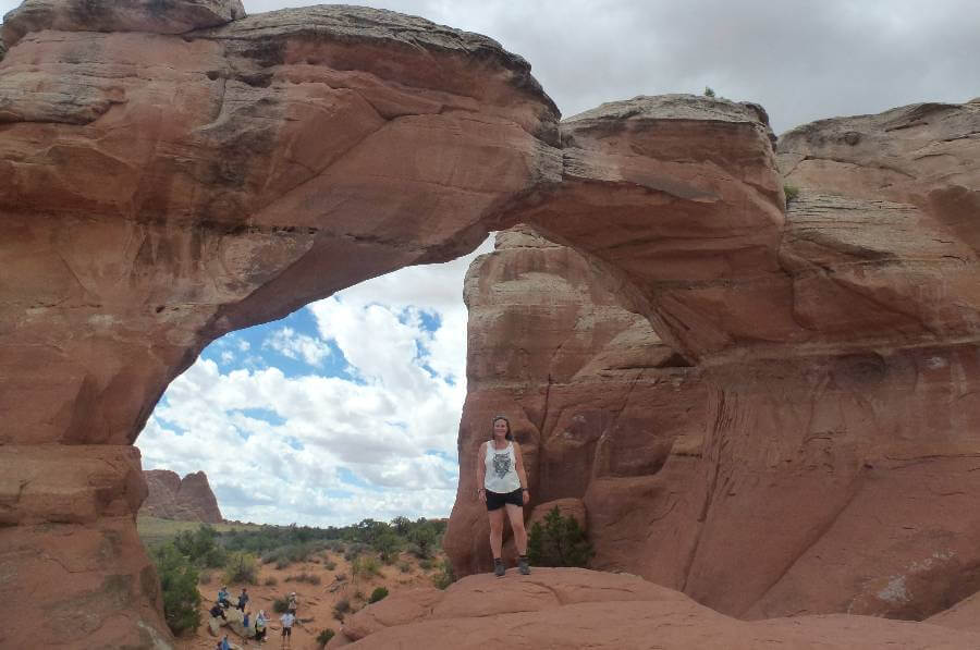 a girl in shorts and tshirt stood in front of an archway through a huge orange rock at arches national park