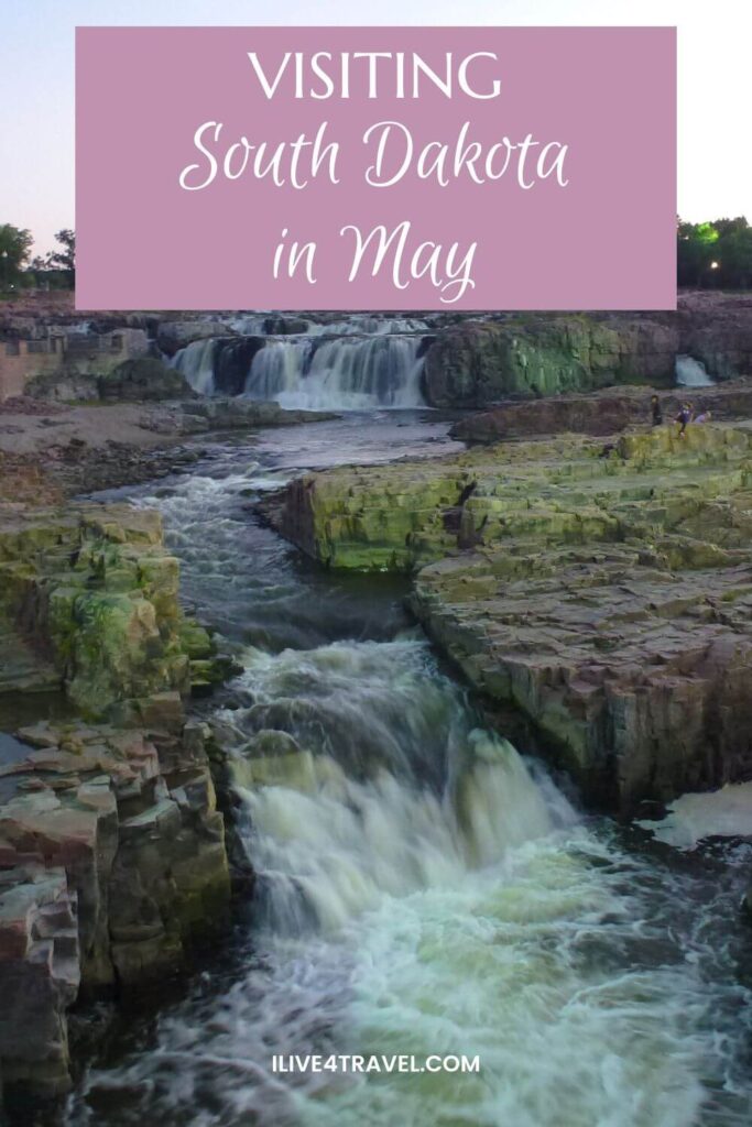 Pinterest pin showing a scenic waterfall in South Dakota during May, with rushing water cascading through layered rock formations and a river cutting through the canyon below. The image includes a soft pink text overlay reading “Visiting South Dakota in May”.