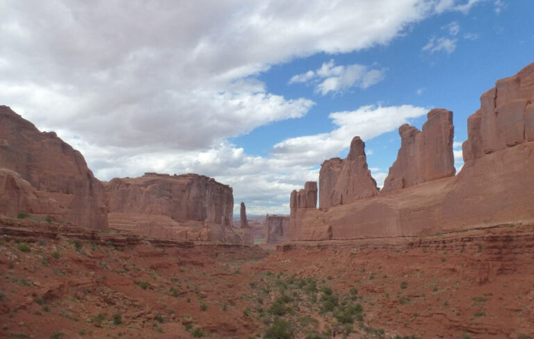 a flat valley with rock towers to the right and in front that have had shapes carved through them over millions of years, the rocks and ground are orange