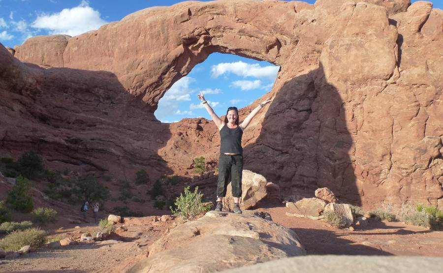 a girl with black trousers and black tshirt stood in front of a large rock with a hole in it, the rock and ground is an orange color