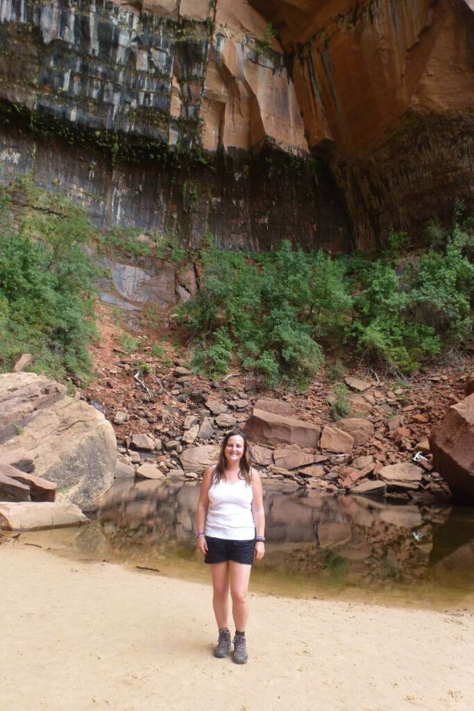 a girl in a white tshirt and black shorts stood in front of a small pool with a orange cliff behind her
