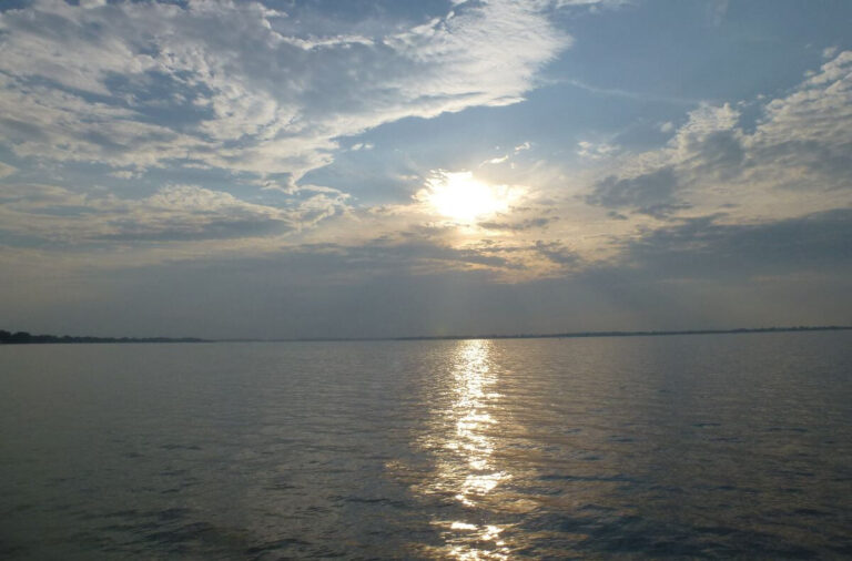 Calm lake scene at sunset in South Dakota, with the sun partially hidden behind layered clouds and its golden reflection stretching across gently rippling water. The distant shoreline sits low on the horizon, creating a peaceful, open landscape under a softly lit evening sky.