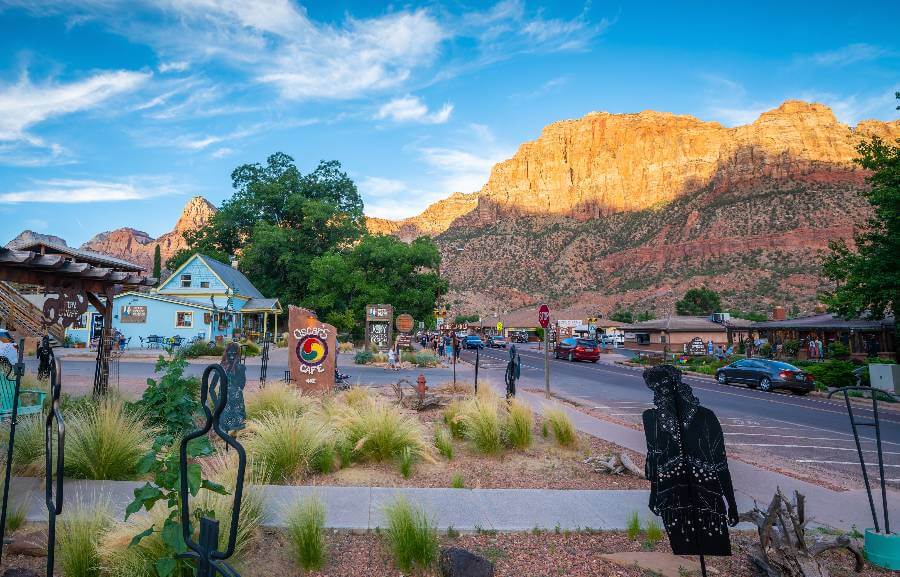 a street through a town, with some metal artwork, buildings alongside the road and in the distance a large orange canyon wall with bushes down the slopes of it