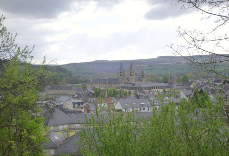 An elevated view over Echternach, Luxembourg, with a historic abbey and twin church towers at the center, surrounded by a cluster of village houses and rolling green hills in the background.