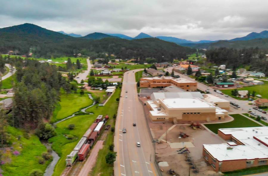 Aerial view of Hill City, South Dakota, showing a main road running through town with cars driving along it. To the left, a railway track with red train cars runs beside a small creek and grassy green fields. Buildings with flat white rooftops and residential houses spread out across the town, surrounded by dense pine forests and rolling hills under a cloudy sky.
