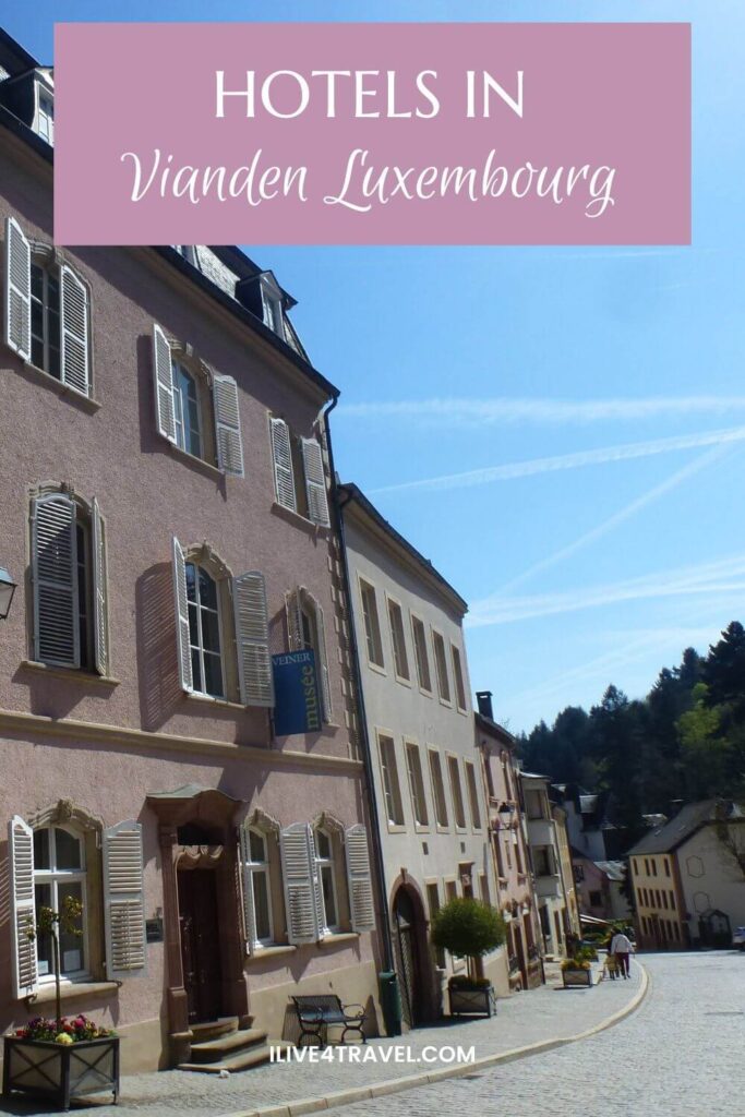Pinterest pin titled “Hotels in Vianden Luxembourg” featuring pastel historic buildings with white shutters lining a quiet cobblestone street in Vianden, Luxembourg, set against a bright blue sky and green hillside in the background.