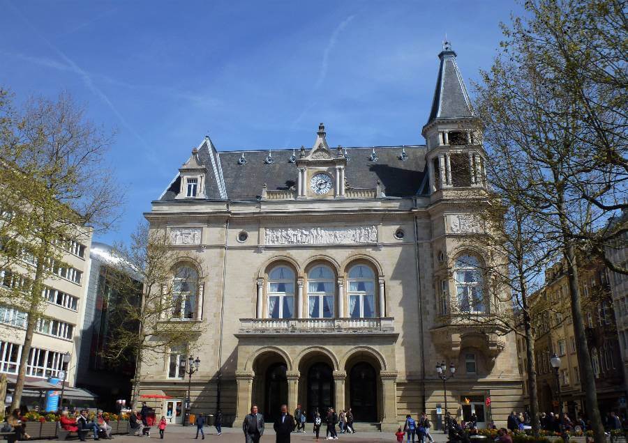 The grand neoclassical facade of Luxembourg City Hall (Cercle Municipal) on a sunny day, viewed from the busy Place d'Armes square. The ornate sandstone building features three arched ground-floor entrances, tall arched windows on the upper floor with a decorative frieze and a central clock, and a steep slate mansard roof with dormer windows. A pointed corner tower rises on the right side. Bare early-spring trees frame the building, and pedestrians mill about the open cobblestone plaza in front.