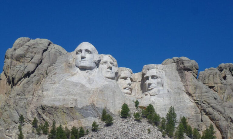 Mount Rushmore National Memorial featuring the carved granite faces of four U.S. presidents—George Washington, Thomas Jefferson, Theodore Roosevelt, and Abraham Lincoln—set against a clear blue sky, with pine trees scattered below the sculpture.
