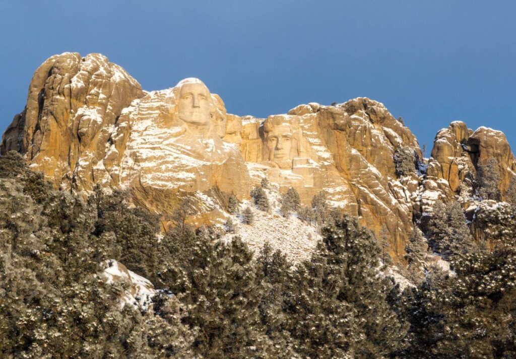 four faces carved into the side of a mountain with trees below them and a sprinkling of 