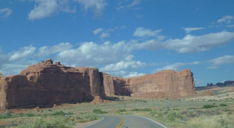 a road driving to a huge area of rock sticking up from the ground, it is orange and alongside the road are a few bushes