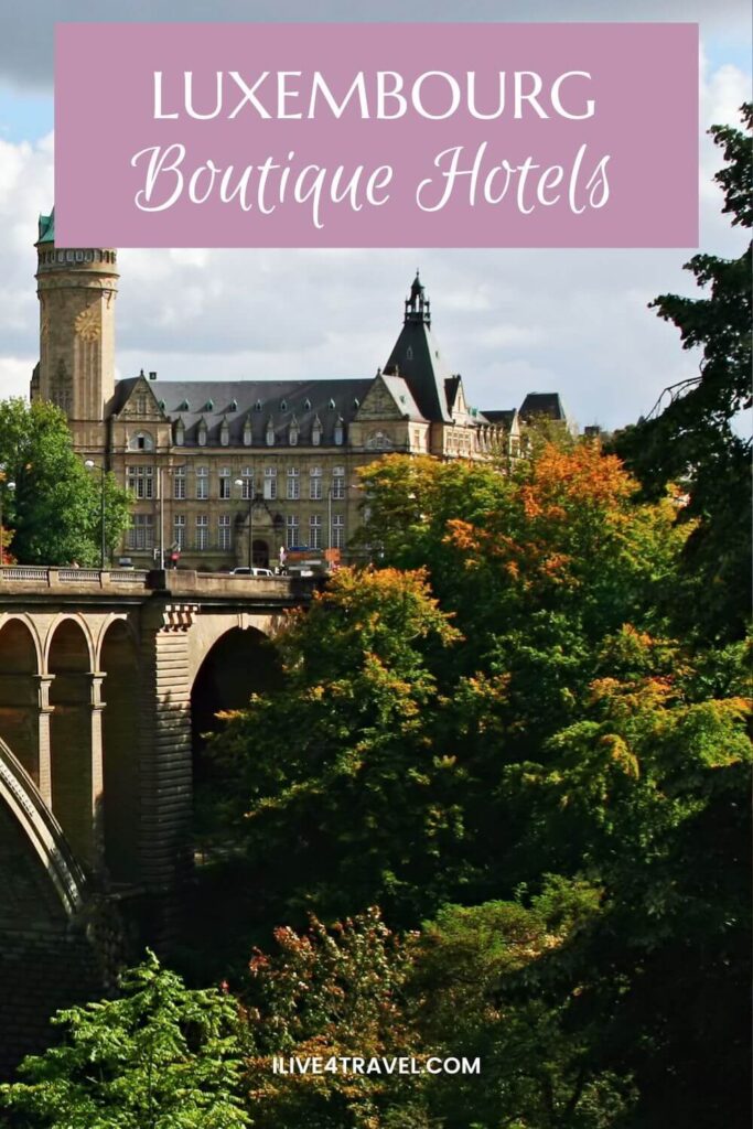 A Pinterest pin titled 'Luxembourg Boutique Hotels'. The image on the pin shows a scenic autumn view of Luxembourg City, with the grand sandstone facade of the Spuerkeess building and its distinctive tower rising above a lush canopy of green and orange-tinged trees. In the foreground, the elegant stone arches of the Adolphe Bridge stretch across the wooded Pétrusse valley below a partly cloudy sky.