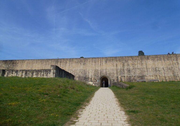 A stone-paved path leads toward an arched entrance in a massive ancient fortress wall built from light-colored sandstone blocks. The tall, imposing wall stretches wide across the frame, with grassy slopes on either side dotted with small yellow wildflowers. A lone figure stands in the shadowed archway. Above, a clear blue sky with faint wispy clouds stretches overhead, and a few trees are visible along the top of the fortification.