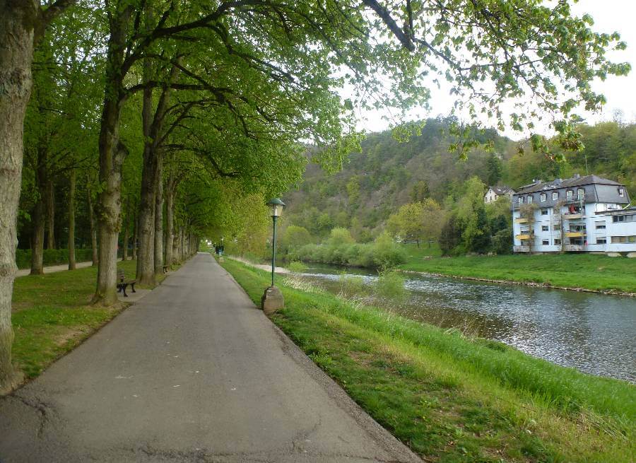Tree-lined walking path along a river in Echternach, Luxembourg, with benches and lamp posts on one side and a gently flowing river on the other, while hillside homes and lush green forest create the scenic backdrop.