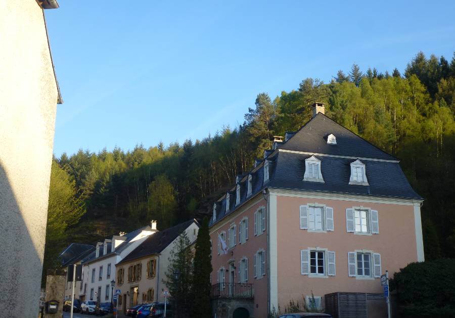 Pastel-colored historic buildings along a quiet street in Vianden, Luxembourg, including a pink building with white shutters and a dark slate roof, set at the base of a steep, forested hillside under a clear blue sky.