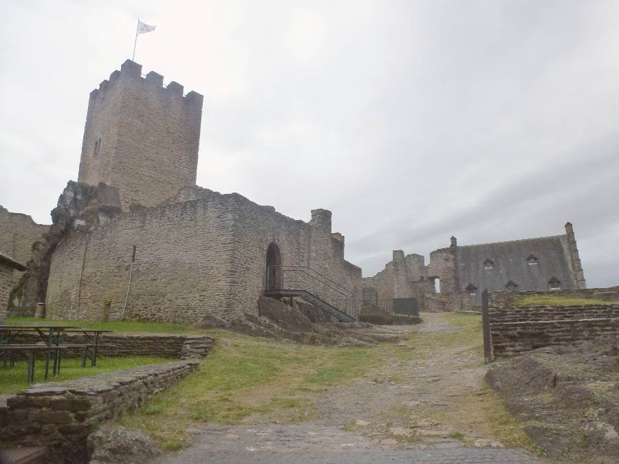 Ruins of a historic stone castle on a hill under an overcast sky, featuring a square tower with a flag on top, crumbling walls, and a narrow path leading up through grassy terrain and stone terraces.