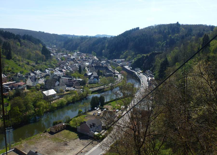 a river running through a town with a road to the right and buildings to the left of the river and the area is surrounded by hills with trees