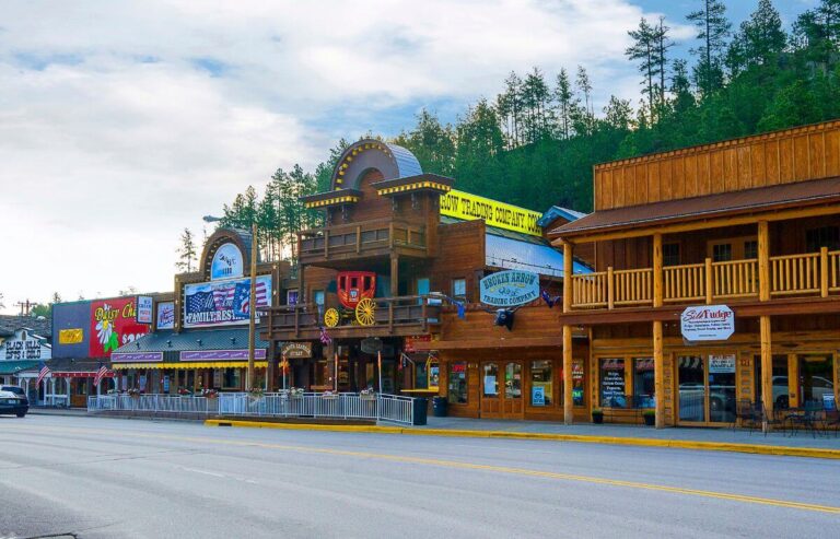 Colorful downtown street in Keystone, South Dakota, with wooden storefronts, Western-style architecture, and a bright red stagecoach display above one building. Shops, restaurants, and signs line the street, with forested hills rising in the background under a partly cloudy sky.