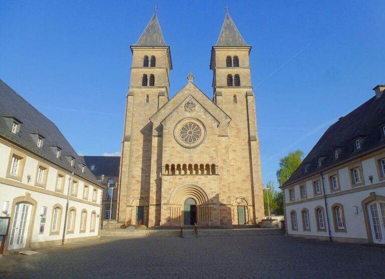 the front of a church in a beige stone with a bell tower at either side of the main door, set in a courtyard with buildings at either side
