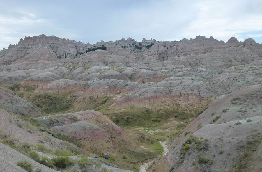 Panoramic view of Badlands National Park in South Dakota, featuring rolling, eroded hills with layered bands of tan, pink, and gray rock, sparse vegetation in the valleys, and jagged ridgelines stretching across the horizon under an overcast sky.