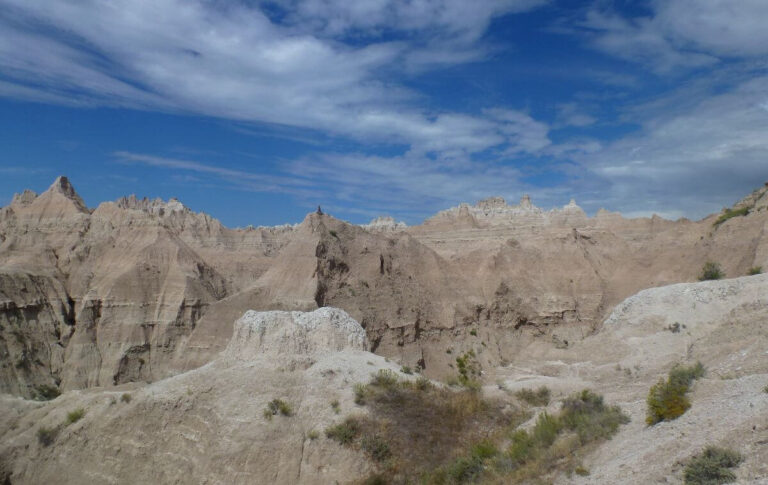 View of Badlands National Park in South Dakota, showing rugged, layered rock formations and sharp spires rising from pale, eroded hills beneath a blue sky with scattered clouds