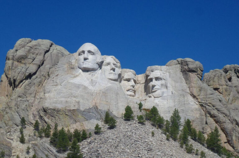 Mount Rushmore National Memorial featuring the carved granite faces of four U.S. presidents—George Washington, Thomas Jefferson, Theodore Roosevelt, and Abraham Lincoln—set against a clear blue sky, with pine trees scattered below the sculpture.
