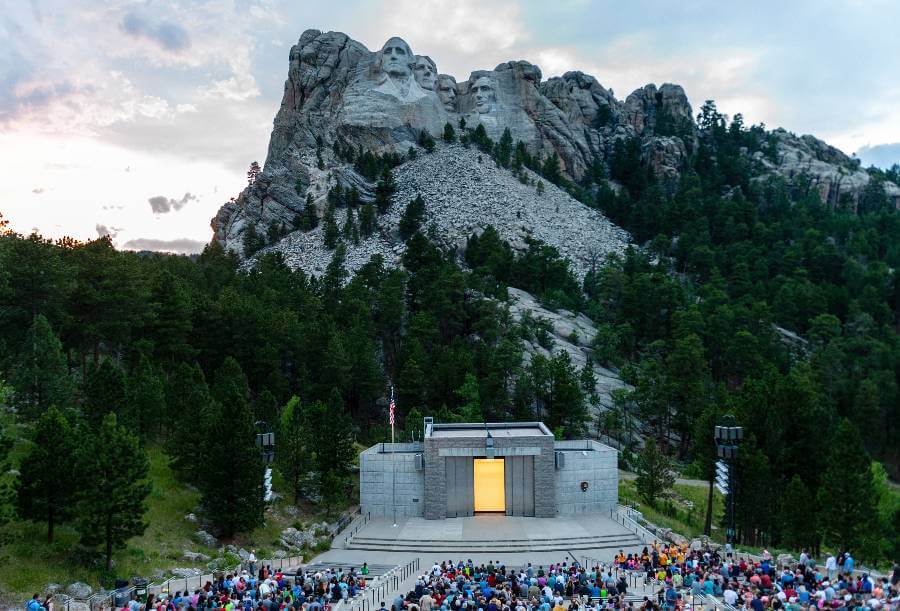 A large crowd gathers at the Mount Rushmore National Memorial amphitheater for the evening lighting ceremony, with the iconic carved faces of George Washington, Thomas Jefferson, Theodore Roosevelt, and Abraham Lincoln visible on the granite cliff in the background, surrounded by dense forest and dramatic skies.